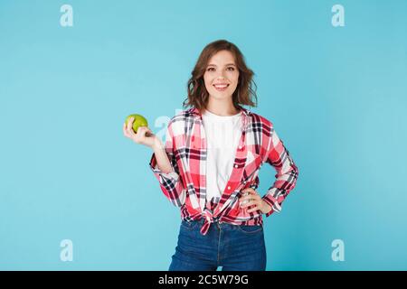 Lächelnd schöne Dame in kariertem Hemd und Jeans halten grünen Apfel, während glücklich in der Kamera über blauen Hintergrund isoliert Stockfoto