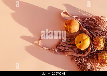 Reife Pfirsiche in Netzbeutel auf rosa Hintergrund mit natürlichen Sonnenstrahlen Schatten, Draufsicht Stockfoto