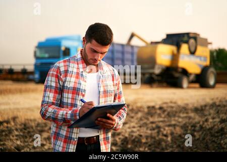 Farmer steuert das Verladen von Weizen vom Erntemaschinen zum Getreidewagen. Fahrer hält Zwischenablage, Notizen, Frachtzählung. Spediteur füllt Sendung aus Stockfoto