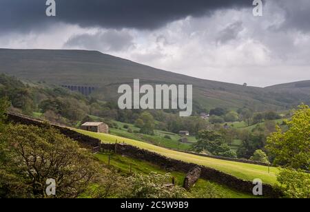 Traditionelle Steinhäuser und Scheunen stehen auf Feldern im Dentdale Tal unter dem Viadukt von Arten Gill auf der Settle-Carlisle Line Bahn und dem Wold Fell Hügel Stockfoto