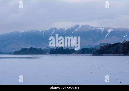 Schnee liegt an den Hängen des Skiddaw Berges über dem gefrorenen See Derwent Water im englischen Lake District. Stockfoto