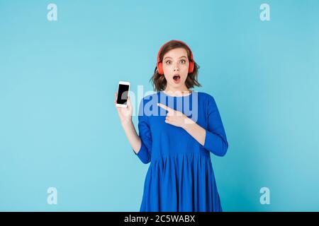 Porträt einer jungen Dame in blauem Kleid und Kopfhörer stehen mit Handy und erstaunt in der Kamera auf über rosa Hintergrund Stockfoto