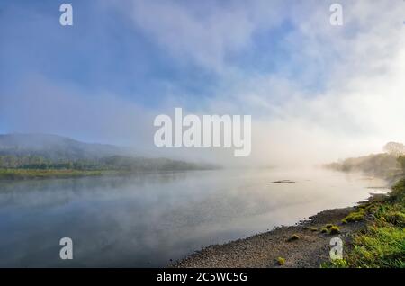 Früher nebliger Morgen über dem Fluss - wunderschöne Sommerlandschaft. Dichter Nebel über der Wasseroberfläche, Spiegelung des Hügels mit Wald bedeckt am Ufer Stockfoto