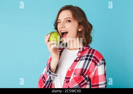 Hübsche Dame im karierten Hemd essen grünen Apfel und glücklich winken Blick in die Kamera über bunten blauen Hintergrund Stockfoto