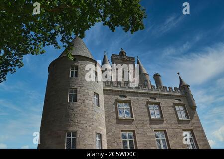 Außenansicht von Glamis Castle in Schottland mit Fenstern und Türmchen, eingerahmt von Laub von Bäumen, mit blauem Himmel und leichten Wolken an sonnigen Tagen Stockfoto