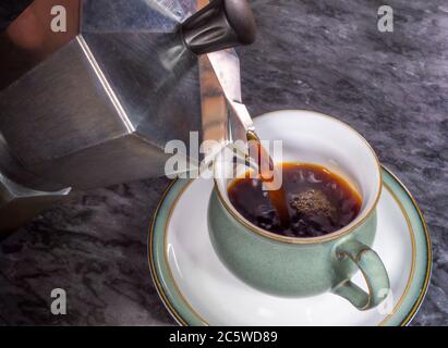 Nahaufnahme von frisch gebrühtem, heißem, dunklem Kaffee, der aus einer traditionellen Ofentopf-Mokakakanne (Perkolator) in eine Tasse in einer Untertasse gießt. Stockfoto