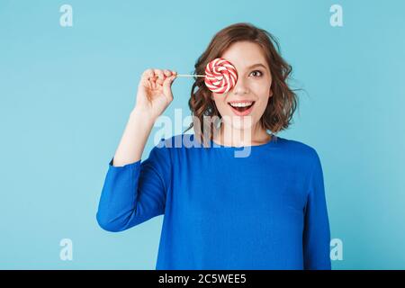 Portrait der schönen Dame im Kleid stehen und ihr Gesicht mit Lutscher Süßigkeiten bedeckt, während glücklich in der Kamera auf blauem Hintergrund suchen Stockfoto