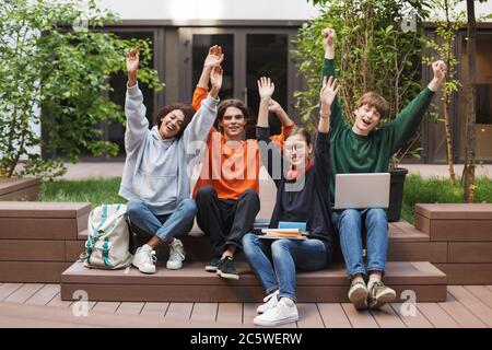Gruppe von fröhlichen Studenten sitzen und glücklich in der Kamera, während sie ihre Hände hoch im Hof der Universität Stockfoto