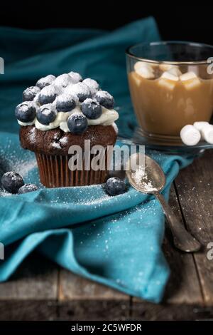 Schokoladenmuffin mit weicher Sahne und frischer Heidelbeere mit Zucker, Tasse Kaffee mit Marshmallows, rustikaler Hintergrund gepudert Stockfoto