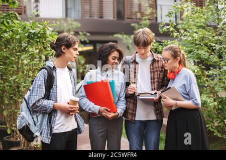 Junge Studenten stehen mit Büchern und Ordnern in den Händen und machen Notizen in Notizbuch, während sie Zeit zusammen im Hof der Universität verbringen Stockfoto