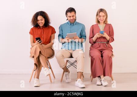 Drei junge interkulturelle Menschen mit modernen mobilen Gadgets und Surfen im Netz, während sie auf Stühlen gegen weiße Wand sitzen Stockfoto