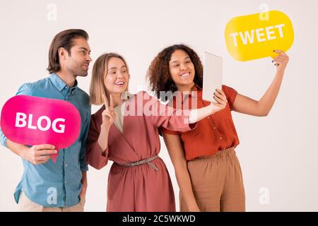 Gruppe von drei fröhlichen freundlichen Millennials machen Selfie, während zwei von ihnen halten Papier Sprechblasen oder Symbole aus sozialen Netzwerken Stockfoto