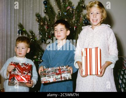 Drei kleine Kinder, Geschwister, bestehend aus einem 7-jährigen Mädchen, einem 5-jährigen Jungen und einem 3-jährigen Jungen, in Schlafanzug und Morgenmänteln, die in den 1960er Jahren in den USA am Weihnachtsmorgen vor einem Weihnachtsbaum Weihnachtsgeschenke halten Stockfoto