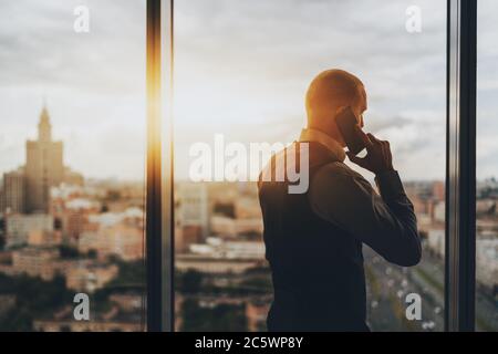 Silhouette des kaukasischen Mannes Unternehmer mit einem Telefongespräch, während neben einem Panoramafenster eines Wolkenkratzers stehen Stockfoto