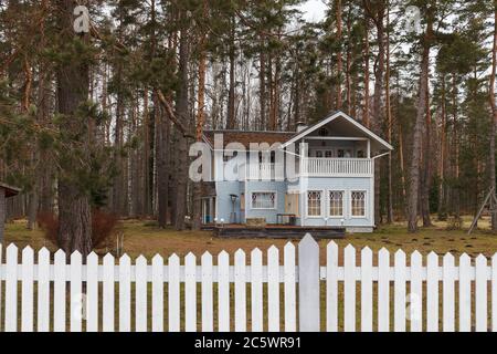 Kleine weiße Holzhütte hinter dem weißen Zaun. Traditioneller nordeuropäischer Stil. Kiefern im Hintergrund. Stockfoto
