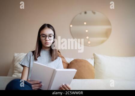 Hübsches Teenager-Mädchen, das zu Hause Buch liest, während es auf dem Sofa sitzt Stockfoto