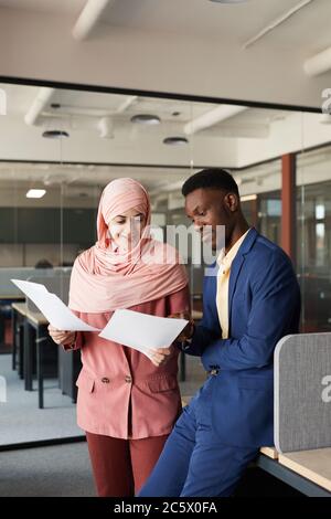 Vertikales Porträt einer modernen muslimischen Geschäftsfrau, die mit afroamerikanischen Kollegen spricht und Dokumente diskutiert, während sie im Büroinneren steht Stockfoto