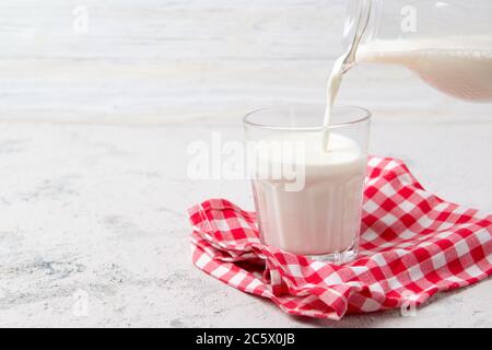 Gießen Sie frische Milch aus dem Krug in ein Glas auf einem roten karierten Handtuch, heller Hintergrund Stockfoto