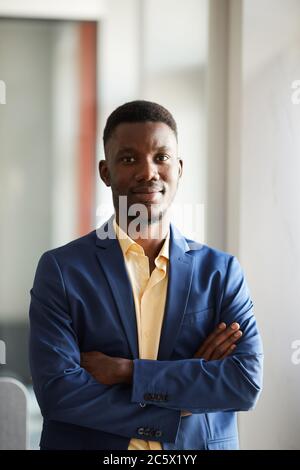 Vertikale Taille bis Porträt des eleganten afrikanischen Geschäftsmann Blick auf Kamera, während sich selbstbewusst stehend mit Armen gekreuzt in modernen Büro Inter Stockfoto