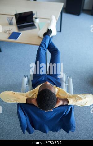 Vertikales Porträt des zeitgenössischen afroamerikanischen Geschäftsmannes, der am Arbeitsplatz im Bürostuhl entspannt, Draufsicht Stockfoto