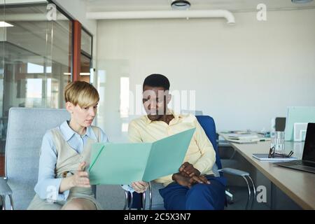 Porträt einer jungen Geschäftsfrau, die Dokumente betrachtet, während sie mit afroamerikanischen Kollegen im Büro über das Arbeitsprojekt diskutiert, Kopierraum Stockfoto