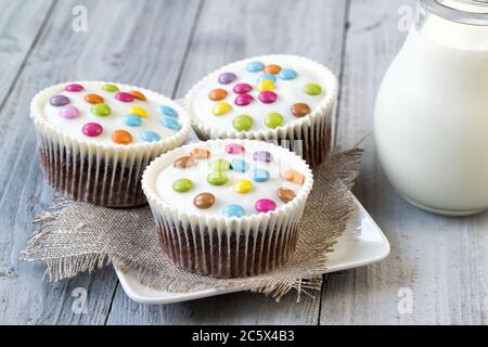 Schokoladenkuchen mit weißer Glasur und bunten Smarties auf einem Teller und Glas Milch, Holzhintergrund Stockfoto