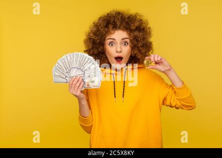 BTC Kryptowährung und Bargeldmittel. Überrascht lockige Frau im urbanen Stil Hoodie hält Dollar-Banknoten, goldene Bitcoin und suchen mit Amazem Stockfoto