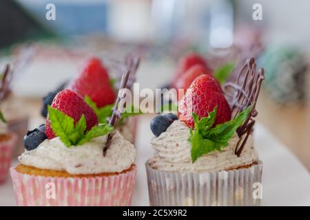 Schöne und bunte Gruppe von Cupcakes mit weißer Peitschencreme, Erdbeeren, Heidelbeeren, Schokolade und Minze auf Holztisch dekoriert. Stockfoto