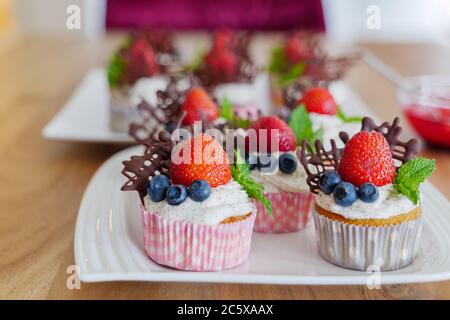 Schöne und bunte Gruppe von Cupcakes mit weißer Peitschencreme, Erdbeeren, Heidelbeeren, Schokolade und Minze auf Holztisch dekoriert. Stockfoto
