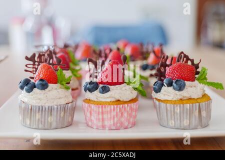 Schöne und bunte Gruppe von Cupcakes mit weißer Peitschencreme, Erdbeeren, Heidelbeeren, Schokolade und Minze auf Holztisch dekoriert. Stockfoto