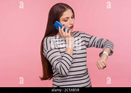 Porträt einer geschäftigen, ungeduldigen, unzufriedenen Frau im gestreiften Sweatshirt, die auf dem Handy spricht und auf die Armbanduhr schaut, die Zeit überprüft, zu m eilt Stockfoto