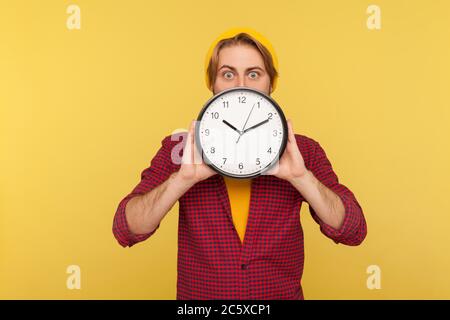 Portrait von staunenden Hipster Kerl in karierten Hemd versteckt Gesicht hinter großen Uhr und Blick auf die Kamera mit besorgt schockiert Ausdruck, zeigt Zeit zu Stockfoto