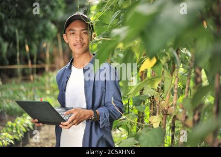 Glücklich von jungen asiatischen Landwirt Mann lächeln, wenn die lange Bohnen Qualität mit Laptop zu überprüfen Stockfoto