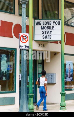 Eine junge afroamerikanische Frau kommt an einem Schild vorbei, das Menschen auffordert, während der Pandemie COVID-19 am 3. Juli 2020 in Mobile, Alabama, in Sicherheit zu bleiben. Stockfoto