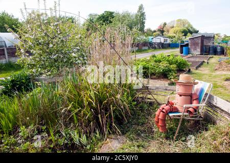 Plantpot Mann auf einem Zuteilungsgebiet, das auf einem Stuhl mit Angelrute an einem überwachsenen Teich sitzt. Die ganze Figur wird aus unterschiedlich großen Pflanztöpfen gemacht Stockfoto