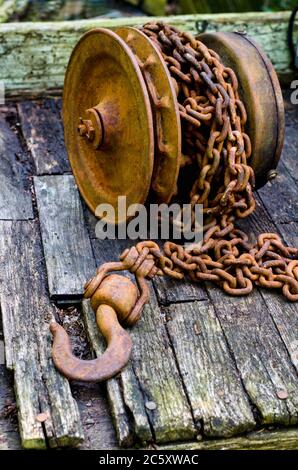 Die verrostete Eisenspule hält eine Länge der schweren Kette mit einem großen rostigen Haken am Ende. Sie liegt auf verrotteten alten Holz Stockfoto