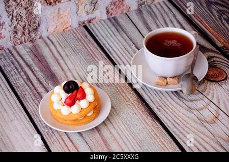 Eine Tasse Tee auf einer Untertasse mit einem Löffel und Rohrzuckerscheiben und einem Teller mit Honigkuchen mit Sahne, Kirsche und Keule. Nahaufnahme. Stockfoto