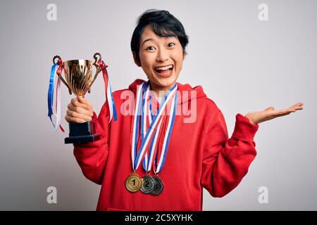 Junge schöne asiatische Mädchen Gewinner mit Trophäe trägt Medaillen auf weißem Hintergrund sehr glücklich und aufgeregt, Sieger Ausdruck feiert Sieg sc Stockfoto