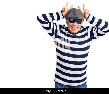 Senior stattlichen Mann trägt Einbrecher Maske und T-Shirt posiert lustig und verrückt mit Fingern auf dem Kopf als Häschen Ohren, lächelnd fröhlich Stockfoto
