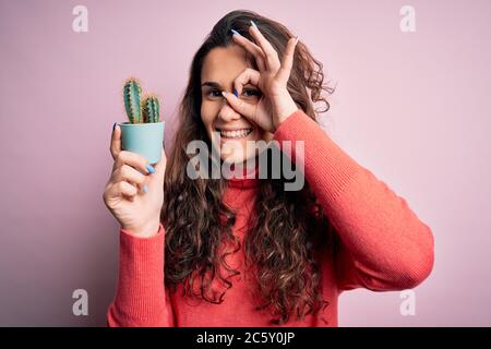 Junge schöne Frau mit lockigem Haar hält kleinen Kaktus über isolierten rosa Hintergrund mit glücklichem Gesicht lächelnd tun ok Zeichen mit Hand auf Auge schauend Stockfoto