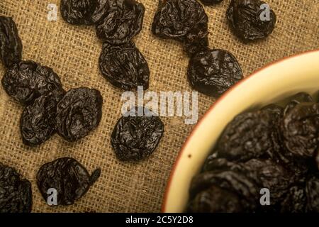 Getrocknete Pflaumen in einer Keramikschale und getrocknete Pflaumen auf einem Hintergrund von grobstrukturiertem Quappen verstreut. Nahaufnahme Stockfoto