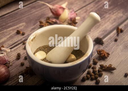 Piment in einem Keramikmörtel und verstreute Knoblauchzehen, Sternanis und Piment auf einem Holztisch. Nahaufnahme Stockfoto