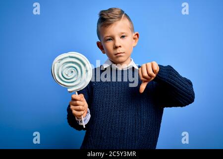 Junge kleine kaukasische Kind essen süße Süßigkeiten Lollipop über blau isoliert Hintergrund mit wütenden Gesicht, negative Zeichen zeigt Abneigung mit Daumen nach unten, Stockfoto