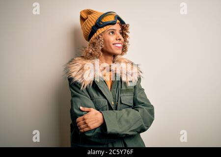 Junge afroamerikanische Skifahrerin Frau mit lockigen Haaren in Schneesportbekleidung und Skibrille, die zur Seite mit gekreuzten Armen schaut, überzeugt und überzeugt Stockfoto