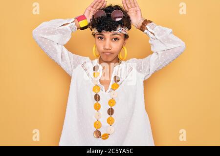 Junge schöne afro amerikanische Hippie Frau trägt Sonnenbrille und Accessoires tun Bunny Ohren Geste mit Händen Handflächen zynisch und sk suchen Stockfoto
