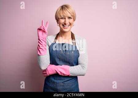 Junge blonde Putzfrau mit kurzen Haaren trägt Schürze und Handschuhe über rosa Hintergrund lächelnd mit glücklichen Gesicht zwinkert an der Kamera tun Sieg s Stockfoto