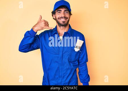 Schöner junger Mann mit lockigen Haaren und tragen Baumeister Jumpsuit Uniform lächelnd dabei Telefongesten mit Hand und Fingern wie auf dem Telefon sprechen Stockfoto