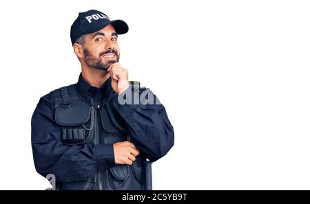 Junger hispanic Mann in Polizeiuniform, der mit gekreuzten Armen und erhobener Hand auf dem Kinn lächelnd auf die Kamera schaut. Positiv denken. Stockfoto