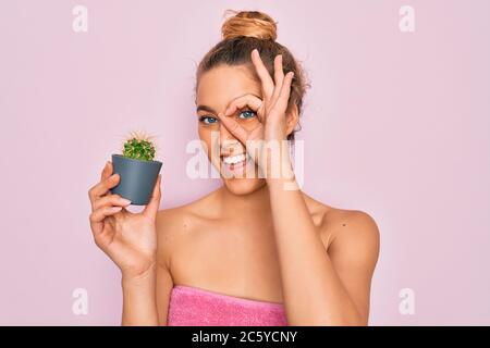 Schöne blonde Frau mit blauen Augen tragen Handtuch Dusche nach Bad mit kleinen Kaktus mit glücklichen Gesicht lächelnd tun ok Zeichen mit Hand auf Auge Blick Stockfoto