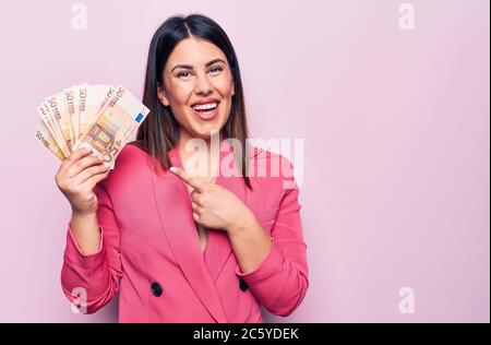 Junge schöne Geschäftsfrau hält Euro-Banknoten über isolierten rosa Hintergrund lächelnd glücklich zeigt mit Hand und Finger Stockfoto